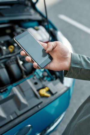 Close up of hand of man holding phone, calling emergency car service while standing near his broken car with open hood on the city streetの写真素材