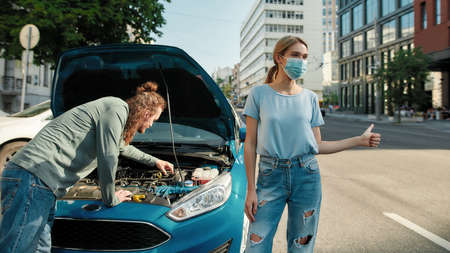 Couple got trouble on the road, Young woman in medical mask showing hitchhiking gesture, asking for help while man examining broken down car, trying to fix itの写真素材