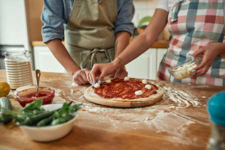 For cheesy taste. Cropped shot of couple making pizza together at home. Man in apron adding, applying tomato sauce on the dough while woman adding mozzarella cheese. Hobby, lifestyleの写真素材
