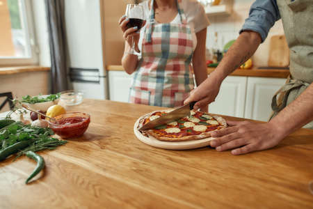 Cropped shot of man, cook cutting hot freshly baked pizza on the table while his girlfriend drinking wine. Couple making pizza together. Hobby, lifestyleの写真素材