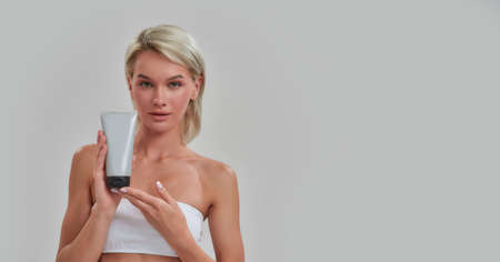 Portrait of young woman with perfect fresh skin looking at camera, holding cosmetic skincare, bodycare product while posing isolated over grey backgroundの写真素材