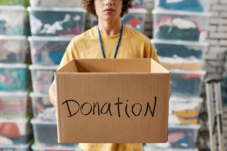 Cropped shot of a guy holding donation box, posing in front of boxes full of clothes, Young male volunteer working for a charity, Focus on cardboard boxの写真素材