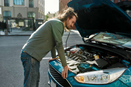 Young man standing near his car with open hood on the city street, looking focused while examining...