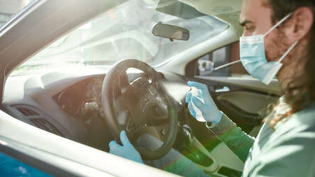 Cropped shot of man wearing medical mask and protective gloves spraying antibacterial disinfectant spray on steering wheel while cleansing car interior to prevent virusの写真素材