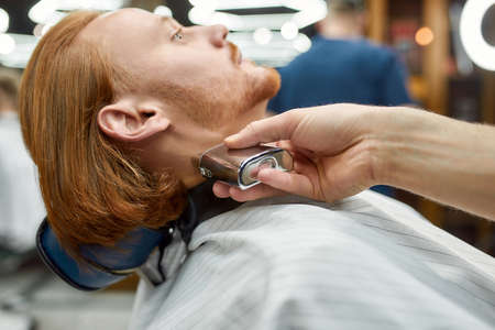 Beard correction. Side view of young redhead guy sitting in barbershop chair getting beard grooming in the modern barbershop. Focus on barber handの写真素材