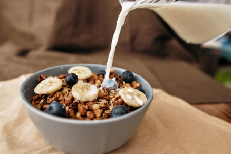 Pouring, adding milk to homemade granola in a plate with nuts, honey, blueberries and banana, served on napkinの写真素材