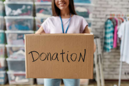 Cropped shot of smiling woman holding donation box, posing in front of rack and boxes full of clothes, Young volunteer working for a charity, Focus on cardboard boxの写真素材