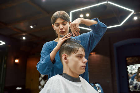 Young professional barber girl or hairstylist with scissors and hair comb in hands making haircut for a man visiting barbershop or beauty salonの写真素材