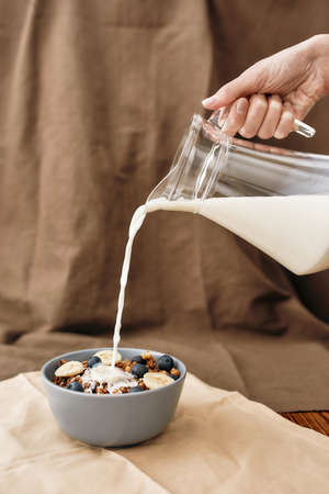 Close up of hand pouring, adding milk to homemade granola in a plate with nuts, honey, berries and...