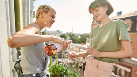 Two women in casual wear pouring wine while standing on the balcony, spending their day togetherの写真素材