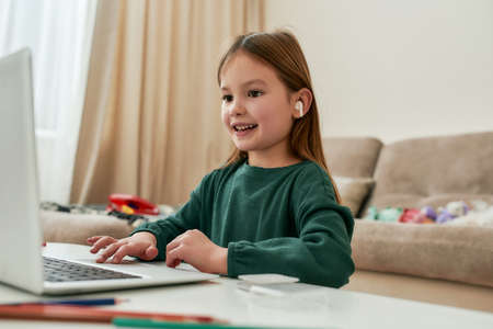 A cute small girl enjoying doing her distance education hometask and smiling while sitting alone in...
