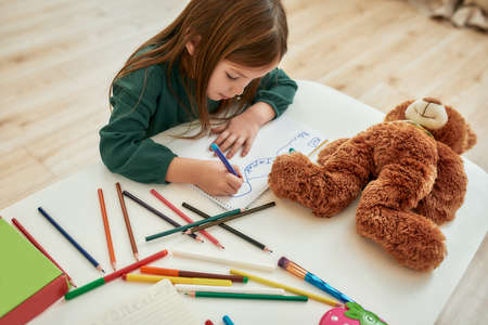 A little girl wearing green sweater sitting alone at the table with a teddy bear drawing with...