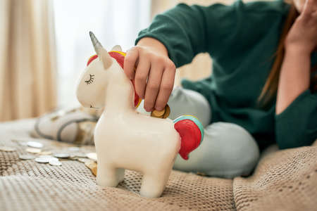 A hand of a small girl is putting a coin into a moneybox while the girl is sitting on a sofa