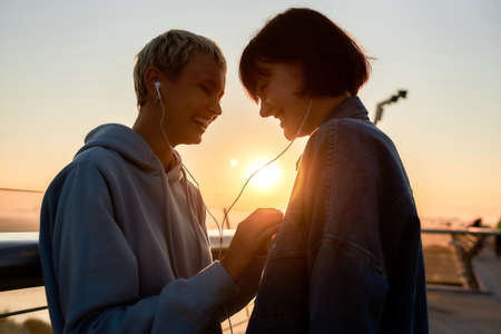 Young lesbian silhouetted couple having fun while listening to music sharing one same earphones, standing on the bridge at sunsetの写真素材
