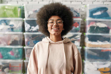 Portrait of happy african american girl smiling at camera while posing in front of boxes full of clothes, Young volunteer working for a charity, donating apparel to needy peopleの写真素材