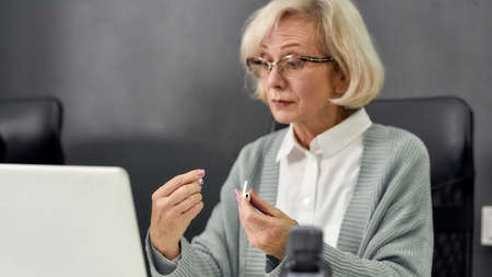 Close up portrait of aged woman, senior intern holding, looking at wireless earphones while using laptop, sitting at desk, working in modern officeの写真素材