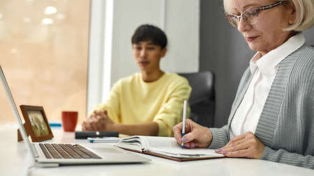 Cropped shot of aged woman, senior intern making notes while listening to her young colleague, Friendly male worker training or teaching new employeeの写真素材