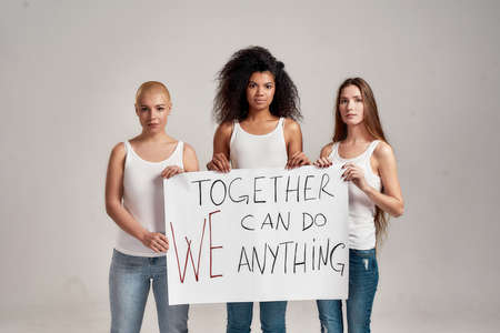 Three confident diverse women in white shirt and casual denim jeans holding, standing with a banner in their hands isolated over grey backgroundの写真素材