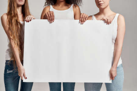 Cropped shot of three diverse women in white shirt holding, standing with a blank banner in their hands isolated over grey backgroundの写真素材