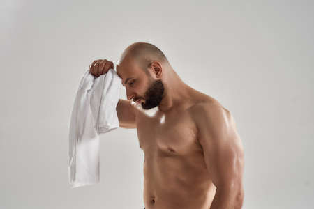Side view of a young muscular caucasian man with naked torso feeling exhausted after hard workout, wiping sweat with white towel while standing isolated over grey backgroundの写真素材