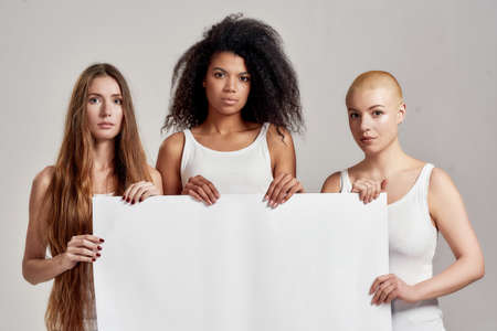 Three confident diverse women in white shirt looking serious at camera while holding, standing with...