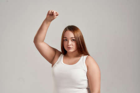 Portrait of a beautiful young caucasian plus size woman wearing white shirt, raising her arm, looking at camera while posing isolated over grey backgroundの写真素材