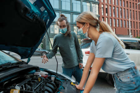 Man in medical mask helping girl with her auto while she is examining, looking at her broken car with open hood, standing on the city streetの写真素材