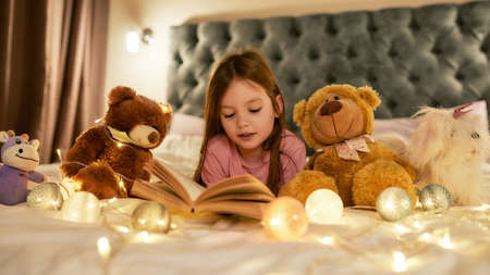 A small girl is lying on her stomack in a big bed barefoot reading a book having a good time with her teddybears and garlands around the bedの写真素材