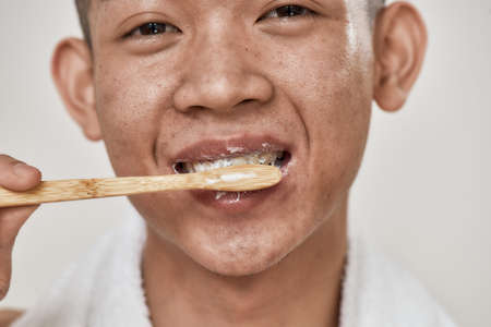 Close up portrait of young asian man with problematic skin brushing his teeth isolated over white...