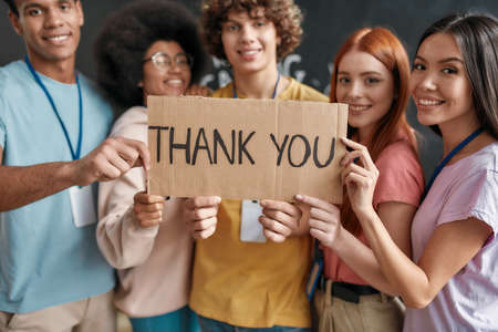 Group of diverse young volunteers smiling at camera, holding card with Thank you lettering while standing in charitable organization officeの写真素材