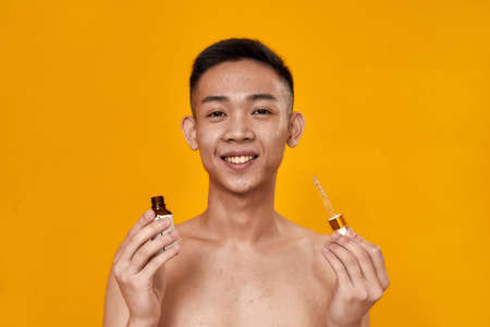 Portrait of young asian man smiling at camera while holding gel serum bottle and glass pipette...