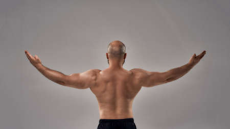 Rear view of muscular strong man with naked torso standing with outstretched arms isolated over grey backgroundの写真素材