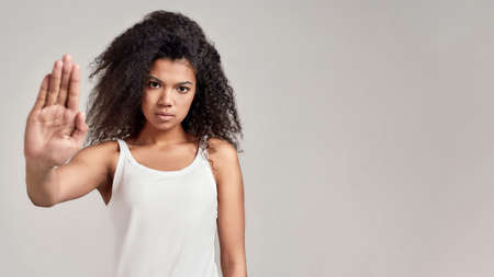 Portrait of young african american woman with curly hair wearing white shirt having serious confident look while showing, making stop gesture isolated over grey backgroundの写真素材