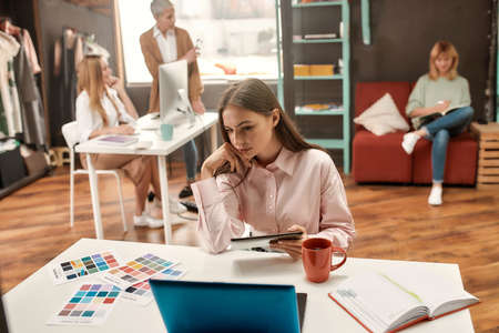 A young and pretty brunette making her decision with a tablet in her hand sitting at a table during a discussion of her female fashionably dressed colleagues on a backgroundの写真素材