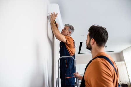 Two workers in uniform, air conditioning masters using ladder while installing a new air conditioner in the apartment. Construction, maintenance and repair conceptの写真素材