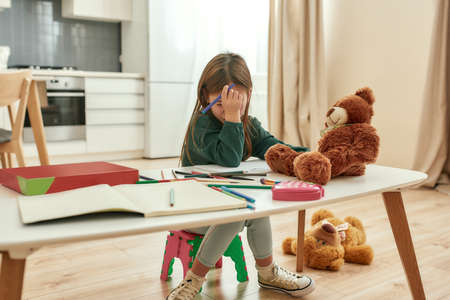 A small girl forgot how to write with her palm over her face sitting at a table in a kitchen with...
