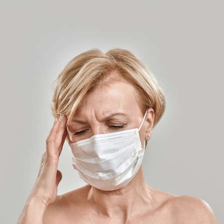 Close up portrait of middle aged woman wearing white medical mask, touching her temples, having...