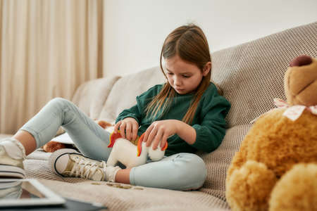 The small girl putting coins into a unicorn moneybox while sitting alone on the sofa in a well lighted room, fully focused on a processの写真素材