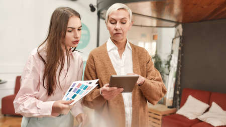 A white mature well-dressed ladyboss holding a tablet giving instructions to a young white...