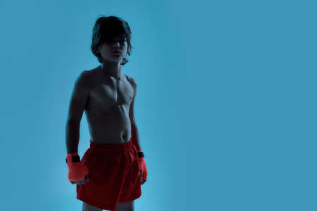 Silhouetted shot of little boy boxer in shorts with boxing bandage on hands, looking at camera while posing, standing isolated over blue backgroundの写真素材
