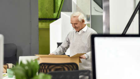 New employee aged man, senior intern looking happy while unpacking box with personal belongings,...