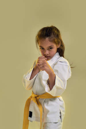 Portrait of little karate girl in white kimono with a yellow sash looking focused at camera,...