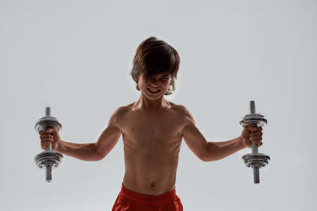 Little sportive boy child with muscular body looking emotional while exercising, lifting weights, standing isolated over grey backgroundの写真素材