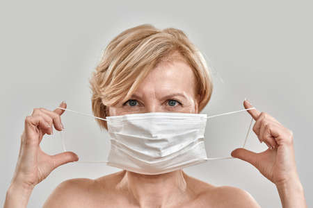 Close up portrait of beautiful middle aged woman holding white medical mask near her face and looking at camera isolated against grey background. Pandemic, safety, prevention conceptの写真素材