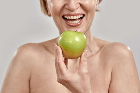 Close up of hand of middle aged woman smiling while holding green apple near her mouth, posing isolated over grey backgroundの写真素材