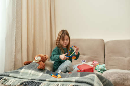 A small girl holding a toy stethoscope while sitting on a sofa with a teddybear and plaid near to...
