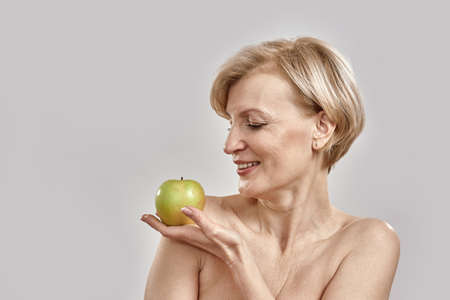 Beautiful middle aged woman holding and looking at green apple, posing isolated over grey backgroundの写真素材