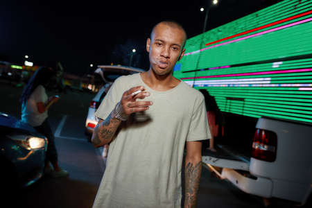 A young dark-skinned casually dressed tattooed man looking into a camera while holding a cigarette...