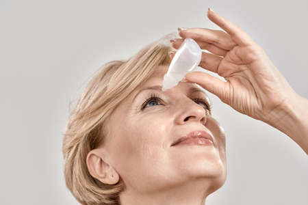 Close up portrait of attractive middle aged woman applying eye drops, standing isolated over grey background