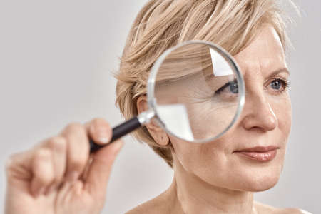 Close up portrait of attractive middle aged woman looking aside, holding a magnifying glass and showing her wrinkles, posing isolated over grey backgroundの写真素材